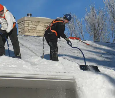 Professional technicians performing roof snow removal with safety harnesses and shovels in ,
